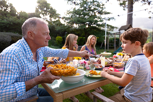 A senior Caucasian man sitting at a table serving his grandson food during a multi-generation family meal in the garden, his relatives serving talking and eating in the background