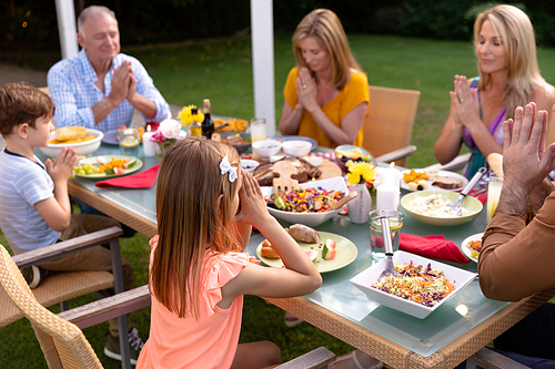 Side view of a Caucasian multi-generation family sitting outside at a dinner table set for a meal, with eyes closed and hands in prayer, saying grace together before eating