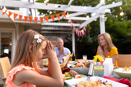 Side view of a Caucasian girl sitting outside at a dinner table set for a family meal, with eyes closed and hands in prayer, saying grace before eating, her grandfather and grandmother background