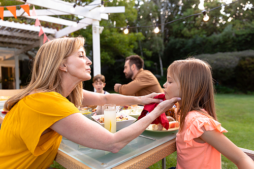 Side view of a Caucasian women sitting in the garden at a dinner table set for a family meal, wiping the fce of her granddaughter with a napkin, the father and brother of the girl in the background