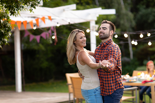Side view of a Caucasian couple in the garden, having fun holding hands, dancing and smiling, their daughter sitting at a table with garden lights on in the background
