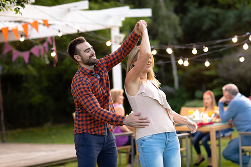 Front view of a Caucasian couple in the garden, having fun holding hands, dancing and smiling, their family sitting at a table with garden lights on in the background