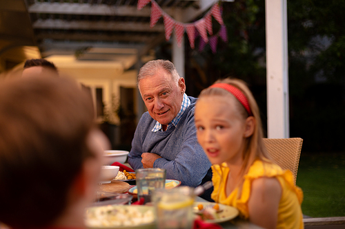 Side view of a senior Caucasian man and his granddaughter sitting at a dinner table for a family celebration meal, looking and listening to another familiy member