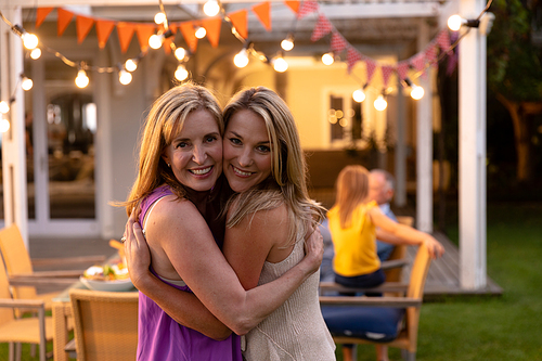 Portrait of a Caucasian woman and her mother standing, embracing and smiling to camera during a multi-generation family celebration meal outside, with members of the family sitting at the dinner table in the background