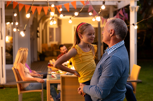 Side view of a senior Caucasian man standing and holding his granddaughter, looking at each other and smiling, during a multi-generation family celebration meal outside with family at the dinner table
