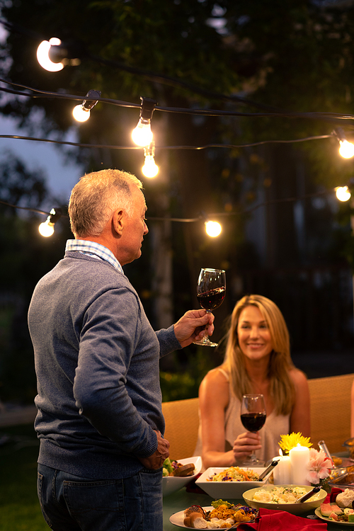 Side view of a senior Caucasian man standing and making a toast holding a glass of red wine, during a family celebration meal outside, his adult daughter sitting at the dinner table and lookng at him