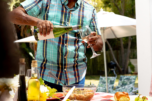Mid section of a Senior African American man spending time in the garden on a sunny day, standing by a table and pouring wine into a glass..