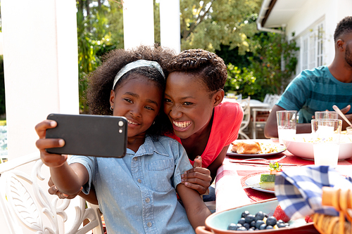 African American family spending time together in the garden on a sunny day, sitting by a table and having a lunch, while a girl man is taking a selfie with her mother with a smartphone.