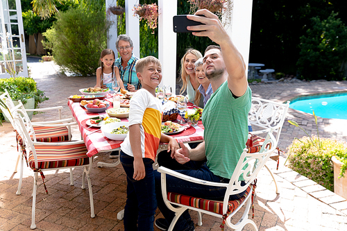 Caucasian three generation family spending time together in the garden on a sunny day, sitting by a table and having a lunch, while a man is taking a selfie with his smartphone.