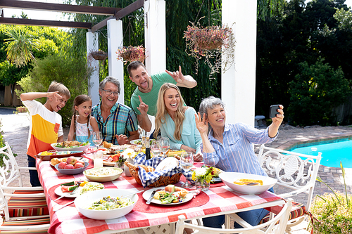 Caucasian three generation family spending time together in the garden on a sunny day, sitting by a table and having a lunch, while a senior woman is taking a selfie with her smartphone.
