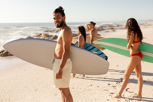 A multi-ethnic group of friends enjoying their time together on a beach on a sunny day, standing, holding surfboards, talking to each other, a mixed race man is looking at the camera