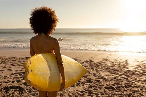 Mixed race woman enjoying her time at a beach with her friends on a sunny day, standing, holding a surfboard and looking at the sea