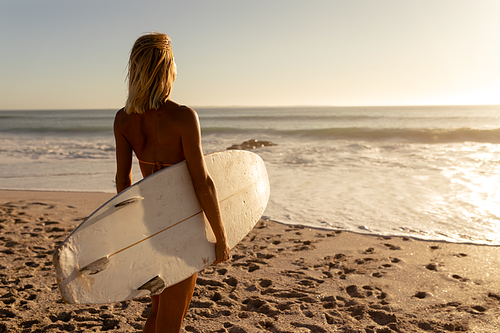 Caucasian woman enjoying her time at a beach with her friends on a sunny day, standing, holding a surfboard and looking at the sea