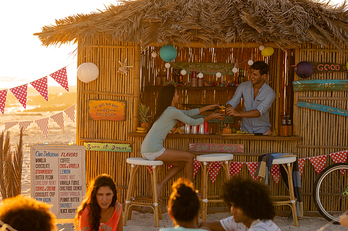 Mixed race woman and Caucasian man enjoying their time on a beach with their freinds during a sunset, sitting by a surfing lessons hut, talking