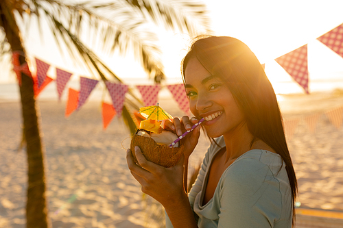 Mixed race woman enjoying her time at a beach with her freinds during sunset, drinking a drink from a coconut, looking at the camera and smiling