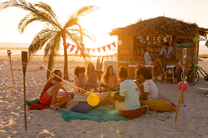 A multi-ethnic group of people enjoying their time at a beach with their friends during a sunset, sitting on sand, playing guitar and smiling