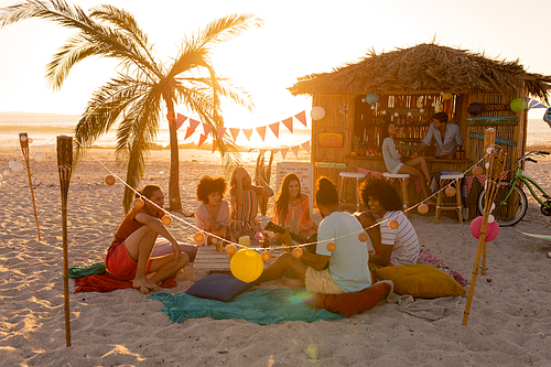 A multi-ethnic group of people enjoying their time at a beach with their friends during a sunset, sitting on sand, playing guitar and smiling