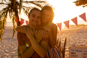 Caucasian man and woman enjoying their time at the beach with their friends during sunset, hugging each other, looking at the camera and smiling