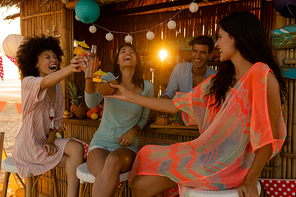 Three mixed race women enjoying their time at a beach with their friends during sunset, sitting by a surfing lessons hut, drinking drinks, making a toast and smiling