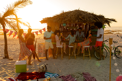 A multi-ethnic group of friends enjoying their time together on a beach during sunset, standing and sitting bu a surfing lessons hut, drinking drinks, talking and smiling