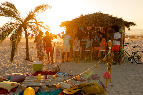 A multi-ethnic group of friends enjoying their time together on a beach during sunset, standing and sitting bu a surfing lessons hut, drinking drinks, talking and smiling