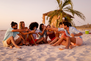 A multi-ethnic group of people enjoying their time at a beach with their friends during a sunset, sitting on sand, drinking drinks and beer, making a toast