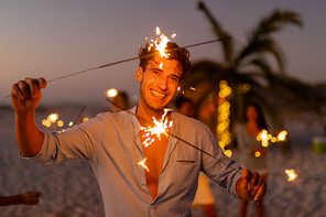 Caucasian man enjoying his time at the beach with his friends during sunset, holding sparklers, looking at the camera and smiling with his friends in the background