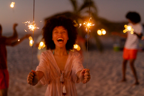 Mixed race woman enjoying her time at the beach with her friends during sunset, holding sparklers, looking at the camera and smiling with her friends in the background