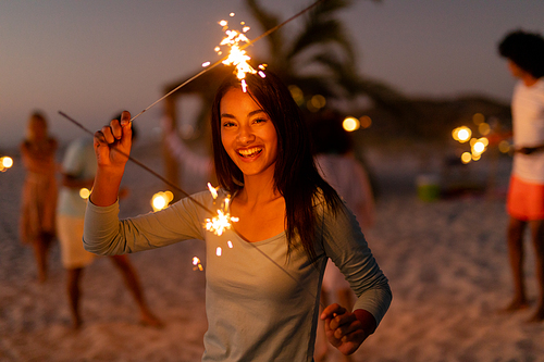 Mixed race woman enjoying her time at the beach with her friends during sunset, holding sparklers, looking at the camera and smiling with her friends in the background