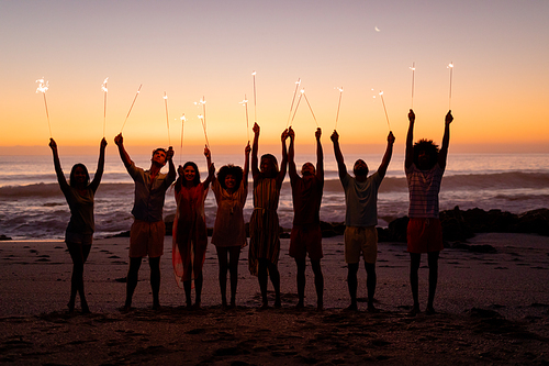 A multi-ethnic group of friends enjoying their time together on a beach during sunset, standing in a row, holding sparklers, raising their arms
