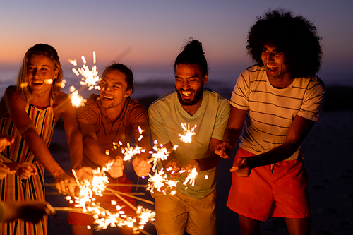 A multi-ethnic group of friends enjoying their time together on a beach during sunset, standing in a circle, holding sparklers and laughing