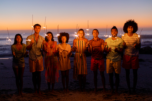 A multi-ethnic group of friends enjoying their time together on a beach during sunset, standing in a row, holding sparklers, looking at the camera and smiling