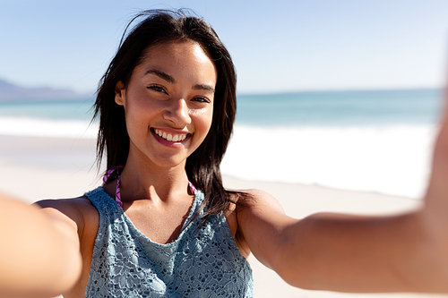 A happy mixed race woman with shoulder length dark hair on holiday, enjoying free time on a sunny beach, smiling and taking a selfie with blue sky and sea in the background