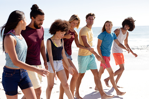 Multi-ethnic group of male and female adult friends wearing shorts and t shirts, walking barefoot on a sunny beach, smiling and talking together, with blue sky and sea in the background
