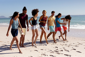 Multi-ethnic group of male and female adult friends wearing shorts and t shirts, walking barefoot on a sunny beach, smiling and talking together, with blue sky and sea in the background