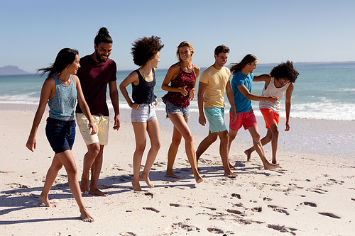 Multi-ethnic group of male and female adult friends wearing shorts and t shirts, walking barefoot on a sunny beach, smiling and talking together, with blue sky and sea in the background