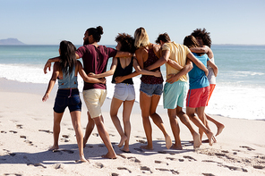 Rear view of multi-ethnic group of male and female adult friends wearing shorts and t shirts, walking barefoot with arms around each other on a sunny beach, with blue sky and sea in the background