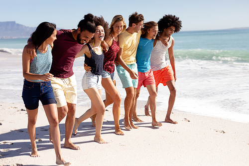 Multi-ethnic group of male and female adult friends wearing shorts and t shirts, walking barefoot with arms around each other on a sunny beach, laughing, with blue sky and sea in the background