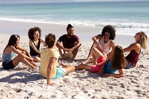 Multi-ethnic group of male and female adult friends wearing shorts and t shirts, sitting on a sunny beach, talking and laughing together, with blue sky and sea in the background