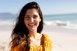 Portrait of a Caucasian woman with long dark hair on holiday, enjoying free time on a sunny beach, looking to camera and smiling, with blue sky and sea in the background