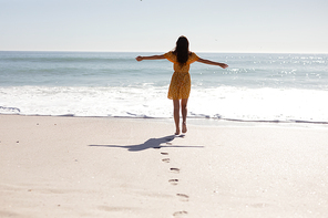 Rear view of Caucasian woman with long dark hair wearing a yellow sundress, walking barefoot on an idyllic sunny beach with her arms in the air, with blue sky and sea in the background