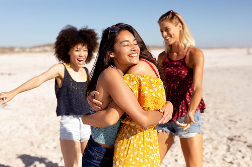 Multi-ethnic group of four female adult friends meeting on a sunny beach, embracing and laughing together, with blue sky and sand in the background