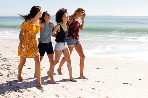 Multi-ethnic group of four female adult friends walking barefoot with arms around each other on a sunny beach, smiling and laughing together, with blue sky and sea in the background