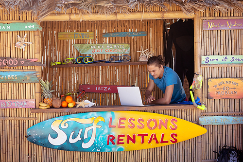 Caucasian man with a hair bun using a laptop on the counter in a beach shack with a sign advertising surf rental and lessons