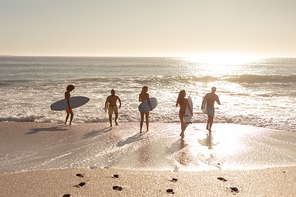 Rear view of a multi-ethnic group of male and female friends on holiday on a beach holding surfboards, running into the sea as the sun goes down