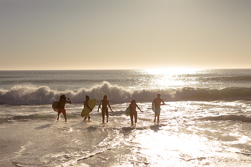Rear view of a multi-ethnic group of male and female friends on holiday on a beach holding surfboards, running into the sea towards a wave as the sun goes down
