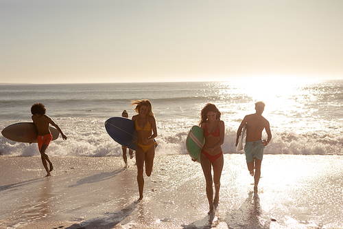 A multi-ethnic group of male and female friends on holiday on a beach holding surfboards, running out of the sea as the sun goes down