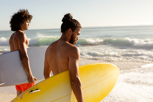 Close up of two Caucasian and mixed race male friends on holiday standing on a sunny beach holding surfboards, looking out to sea and and waiting for wave, with sunny sky and sea in the background