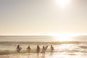 Rear view of a multi-ethnic group of male and female friends on holiday on a beach holding surfboards, wading into the sea as the sun goes down