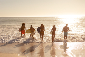 A multi-ethnic group of male and female friends on holiday on a beach holding surfboards, running out of the sea as the sun goes down